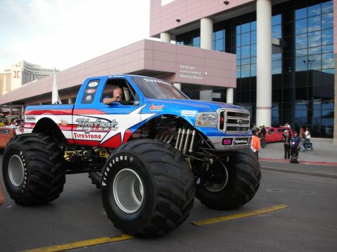 1998 Ford Crown Victoria - blue and white monster truck on enormous Firestone tires rolls along a street outside a large convention building.