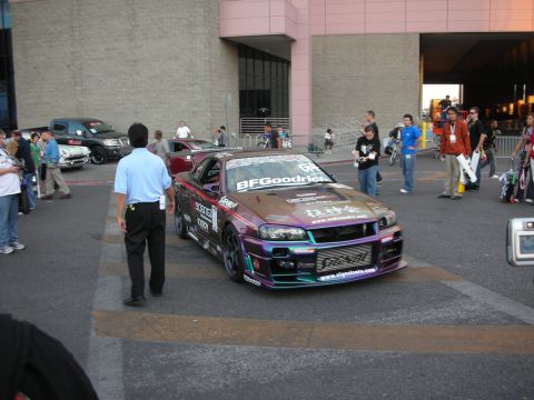 1998 Ford Crown Victoria - purple race-liveried coupe with BFGoodrich and sponsor decals sits among a crowd on a street outside a convention venue.
