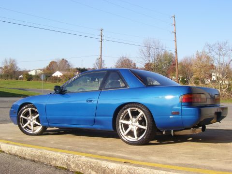 1993 Nissan 240SX - A blue coupe with aftermarket wheels is parked on a concrete surface next to a yellow line.