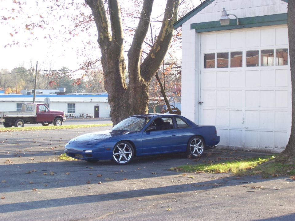 Lowered blue Nissan 240SX hatchback on silver aftermarket wheels parked by a white garage