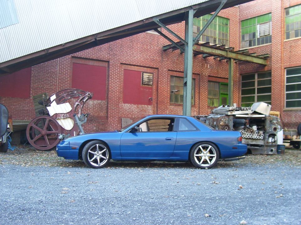 Blue Nissan 240SX coupe lowered on aftermarket wheels, profile view by a brick factory