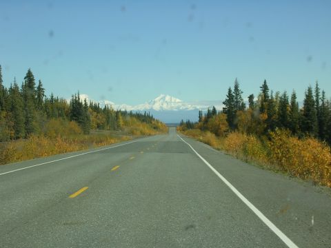 1982 Chevrolet C/K Pickup - A highway stretches towards snow-capped mountains, flanked by autumn foliage and evergreen trees under a clear blue sky.
