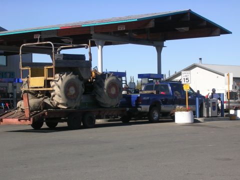 1982 Chevrolet C/K Pickup - A blue pickup truck tows a large tractor on a trailer past gas pumps under a covered canopy.