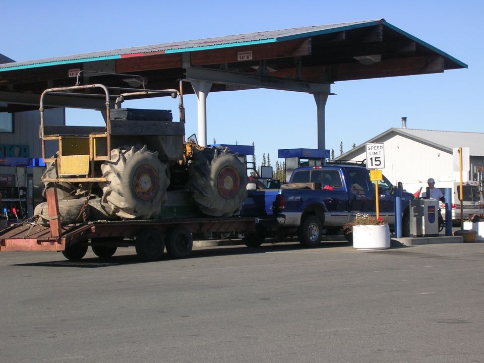 Blue pickup truck towing a large tractor on a trailer at a gas station