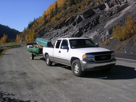 1982 Chevrolet C/K Pickup - A white GMC pickup truck tows a trailer carrying a green canoe along a scenic mountain road.