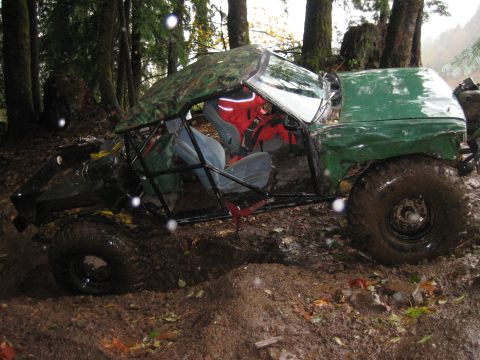 1990 Ford Bronco - This heavily modified off-road vehicle, possibly a Ford Bronco, is deeply stuck in the mud amidst a wooded area.