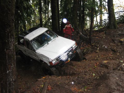 1990 Ford Bronco - A white pickup truck navigates a muddy, wooded trail, its front wheel deep in the mire.