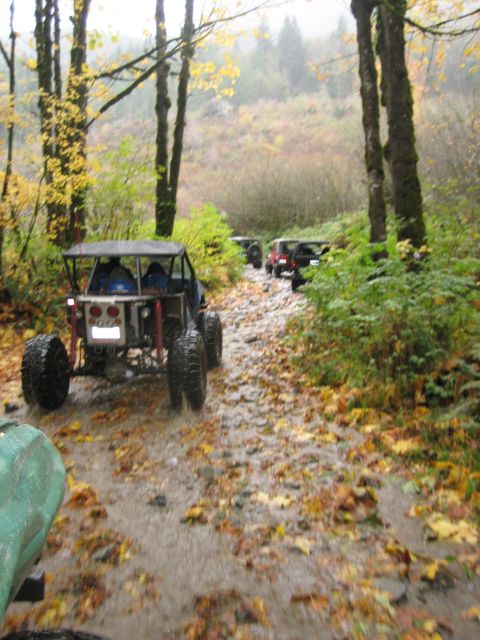 1990 Ford Bronco - Multiple off-road vehicles navigate a wet, wooded trail during autumn.