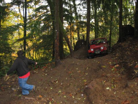 1990 Ford Bronco - A person uses a rope to help pull a red Jeep Wrangler up a steep, wooded dirt trail.