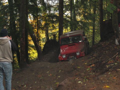 1990 Ford Bronco - A red Jeep Wrangler navigates a challenging muddy trail surrounded by trees.