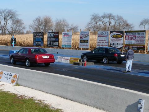 1997 Mercury Grand Marquis - Two sedans race down a drag strip with advertising billboards in the background.