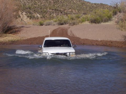1993 Ford Bronco - A white SUV navigates through a water crossing on a dirt path, with arid hills in the background.