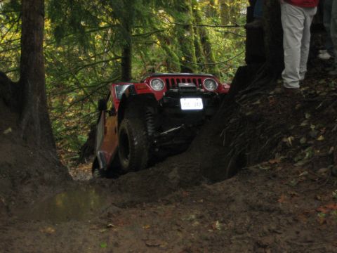 1990 Ford Bronco - A red Jeep Wrangler tackles a challenging muddy off-road course surrounded by dense forest.