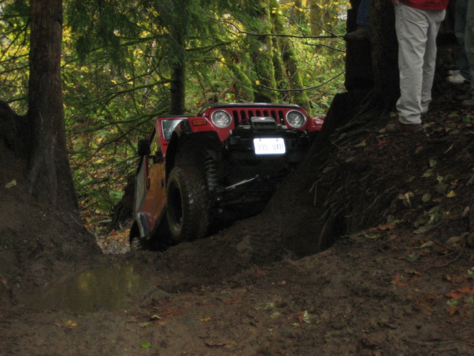 Red Jeep Wrangler navigating a muddy trail between trees, front view with winch visible