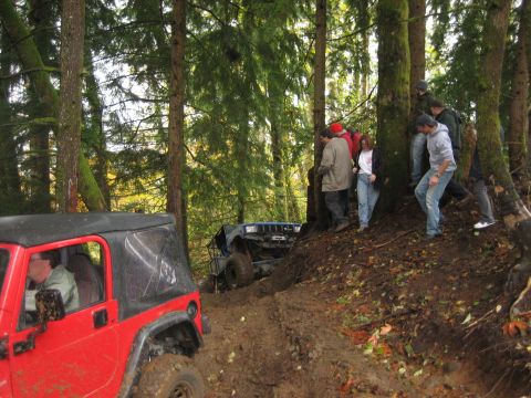 1990 Ford Bronco - Two off-road vehicles navigate a challenging muddy path through a dense forest with onlookers.