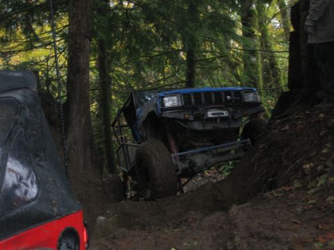 1990 Ford Bronco - Two off-road vehicles navigate a challenging, muddy trail through a dense forest.