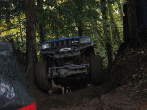 1990 Ford Bronco - An off-road SUV navigates a steep, muddy incline in a wooded area, with another vehicle partially visible to the left.