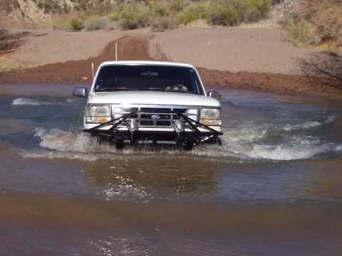 1993 Ford Bronco - A white Ford Bronco navigates a shallow water crossing on a dirt road.