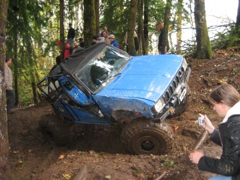 1990 Ford Bronco - A blue off-road vehicle navigates a muddy trail with spectators watching.