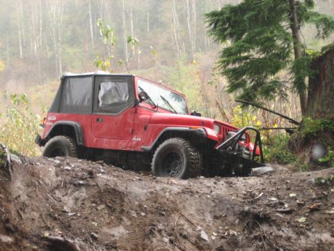 1990 Ford Bronco - A red Jeep Wrangler navigates a challenging muddy off-road trail in a wooded area.