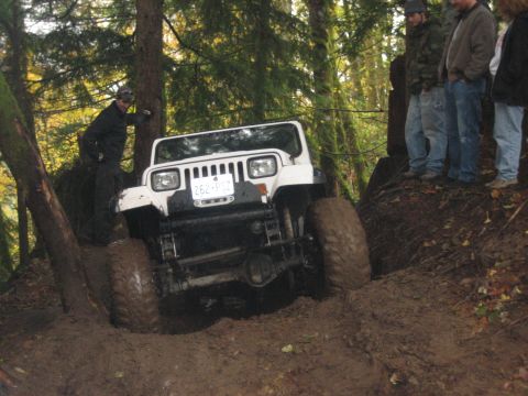 1990 Ford Bronco - A white Jeep Wrangler tackles a challenging muddy trail with onlookers observing.