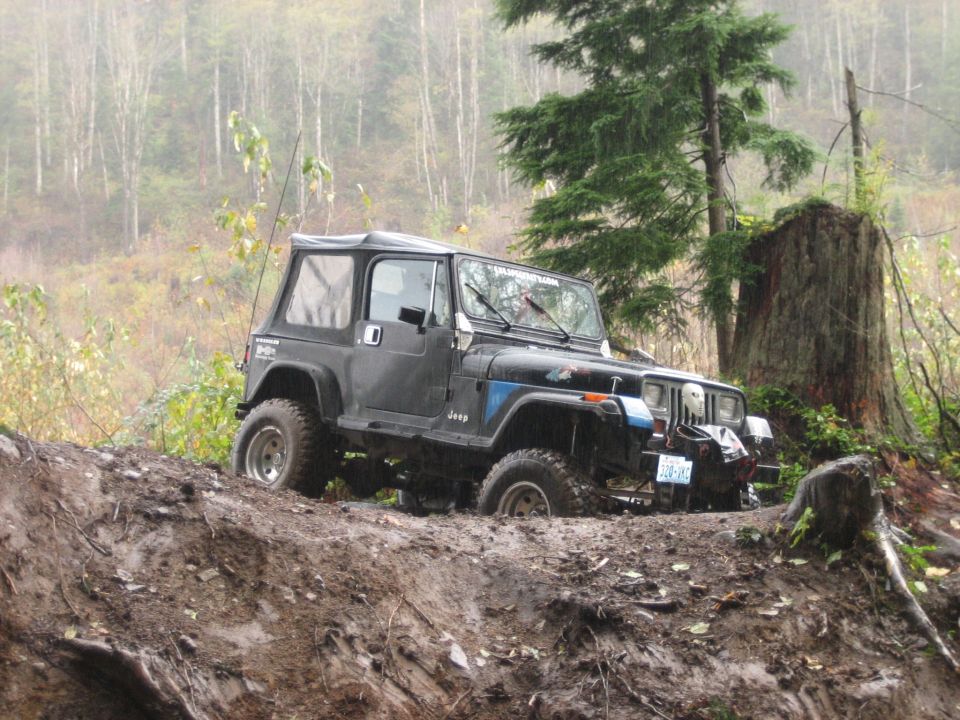 Black Jeep Wrangler with soft top stuck in mud on a wooded trail. The front grille and headlights are visible