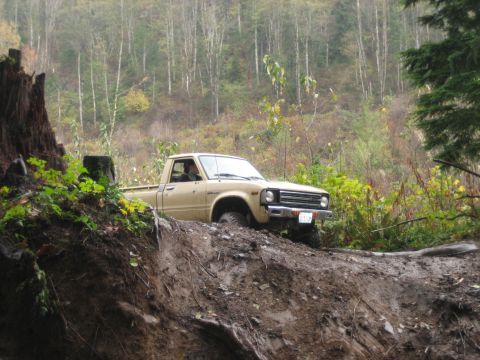 1990 Ford Bronco - A tan pickup truck navigates a steep, muddy incline in a wooded area.