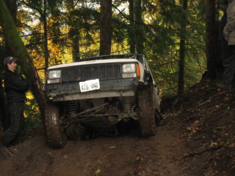 1990 Ford Bronco - A Jeep Cherokee is shown off-roading on a muddy trail surrounded by trees.