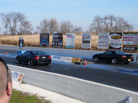 1997 Mercury Grand Marquis - Two black muscle cars compete on a drag strip, with various sponsor signs lining the track.