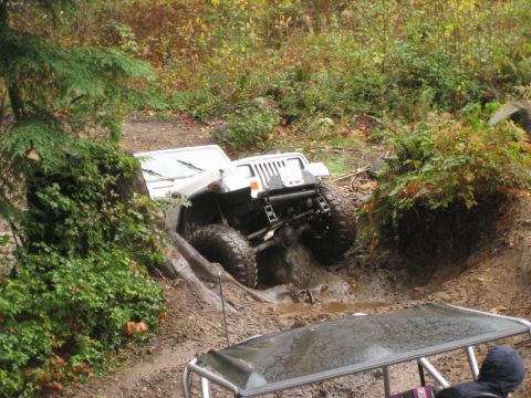 1990 Ford Bronco - White Jeep Wrangler navigating a muddy off-road trail, with another vehicle partially visible.