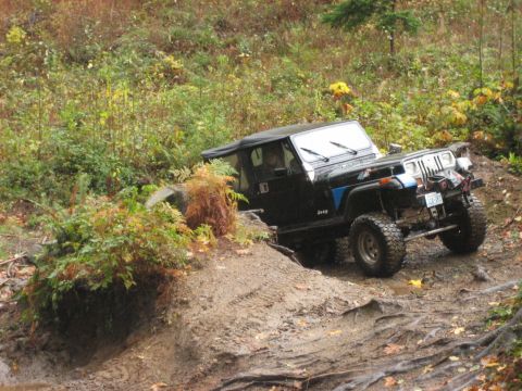 1990 Ford Bronco - A Jeep Wrangler is shown off-roading on a wet, muddy trail surrounded by trees and plants.