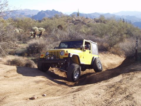 1993 Ford Bronco - A yellow Jeep Wrangler navigates a dusty, off-road trail with desert mountains in the background.