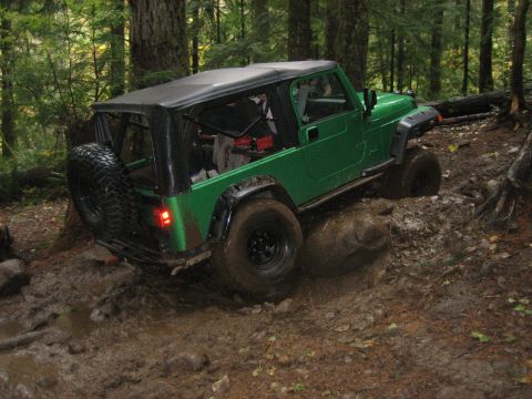 1990 Ford Bronco - Green Jeep Wrangler tackling a challenging muddy trail in a forest setting.