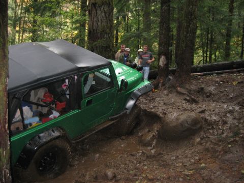 1990 Ford Bronco - A group of people watch as a green Jeep Wrangler navigates a muddy off-road trail.