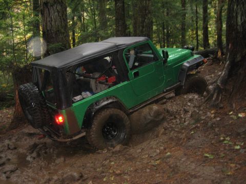 1990 Ford Bronco - A green Jeep Wrangler navigates a muddy path in a dense forest.