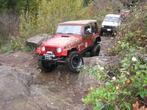 1990 Ford Bronco - A red Jeep Wrangler tackles a challenging off-road course, with other vehicles visible on the trail behind it.