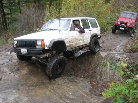 1990 Ford Bronco - A white Jeep Cherokee navigates a challenging rocky path, with a red Jeep Wrangler in the background.