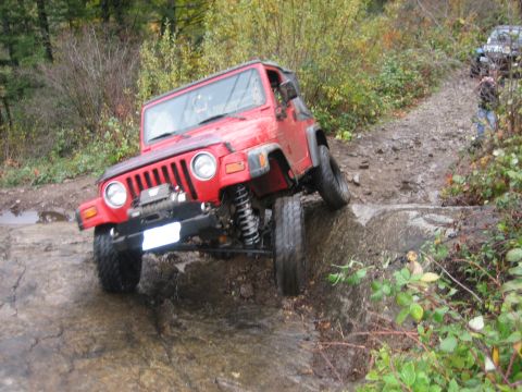 1990 Ford Bronco - A red Jeep Wrangler tackles a challenging off-road course, with a second vehicle visible on the trail behind it.