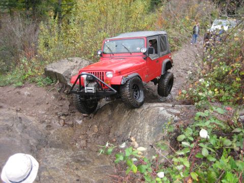 1990 Ford Bronco - A red Jeep Wrangler tackles a challenging rocky off-road course, with another vehicle partially visible further down the path.