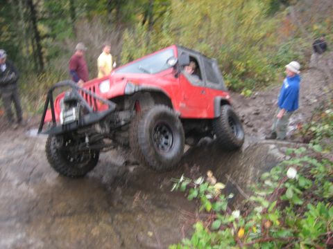 1990 Ford Bronco - A red Jeep Wrangler navigates a muddy off-road trail with onlookers present.