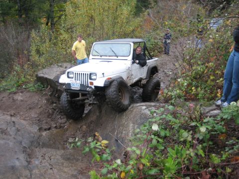 1990 Ford Bronco - White Jeep Wrangler tackling a challenging off-road obstacle on a wooded trail.