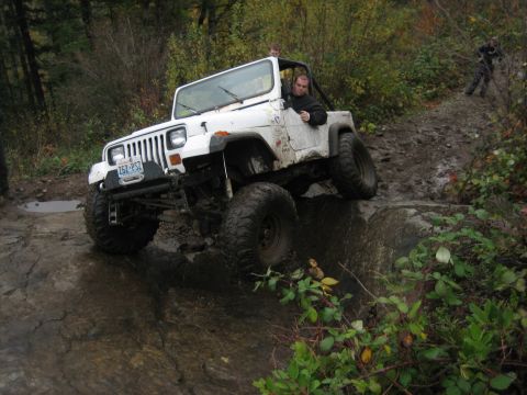 1990 Ford Bronco - A white Jeep Wrangler navigates a challenging off-road course with muddy terrain and large rocks.