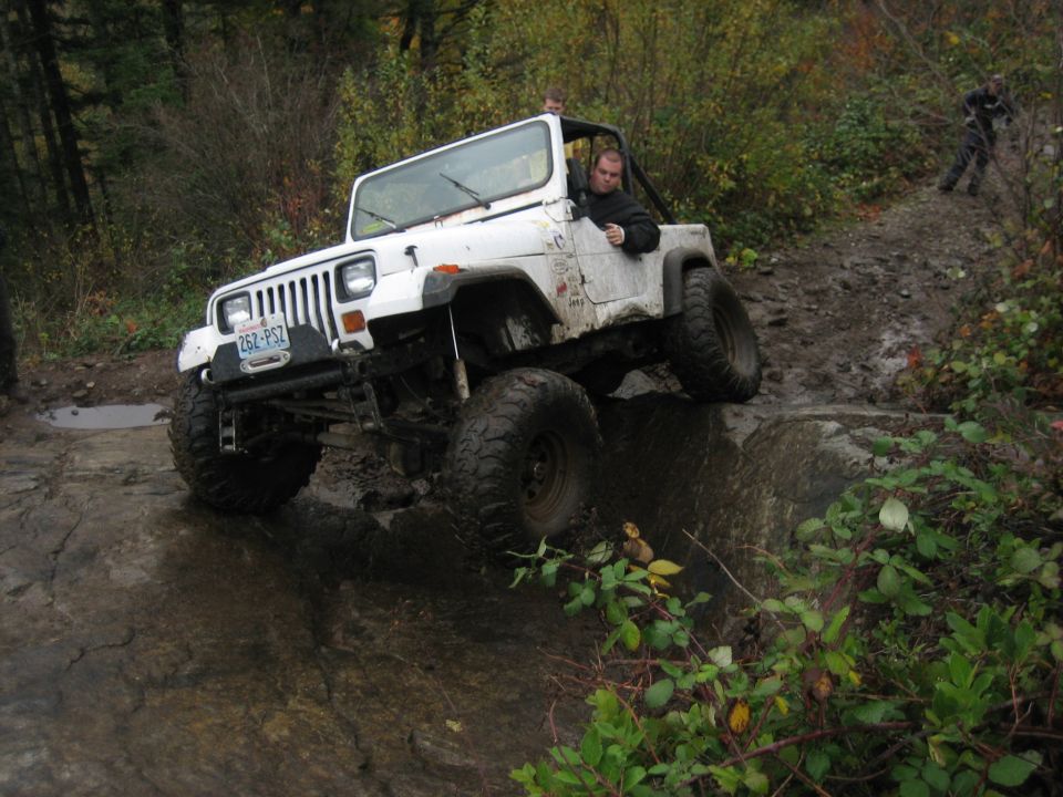 White Jeep Wrangler driving over rocks on a muddy trail. Driver is visible. Another person is in the background