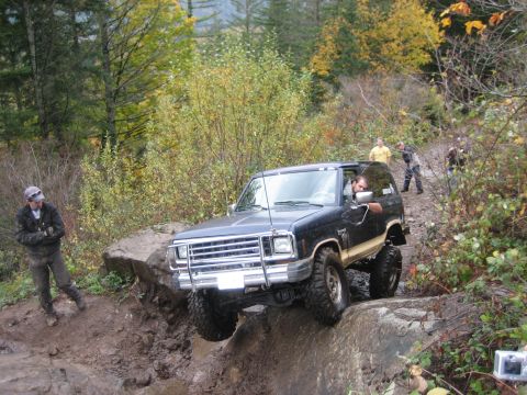 1990 Ford Bronco - A dark-colored Ford Bronco navigates a challenging off-road course with onlookers present.