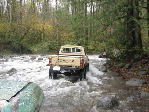 1990 Ford Bronco - Toyota pickup truck navigates a rocky riverbed in a wooded area, with another vehicle following.
