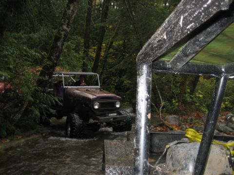 1990 Ford Bronco - Two off-road vehicles navigate a wooded stream, with one vehicle's roll cage prominently in the foreground.