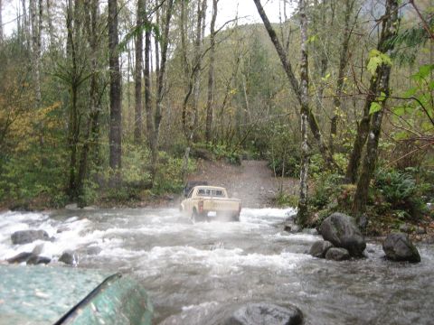 1990 Ford Bronco - A Toyota pickup truck navigates a shallow, rocky river in a wooded area.