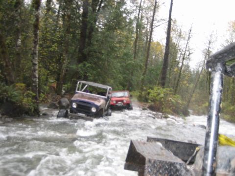 1990 Ford Bronco - Two off-road vehicles are seen navigating a river in a wooded area.