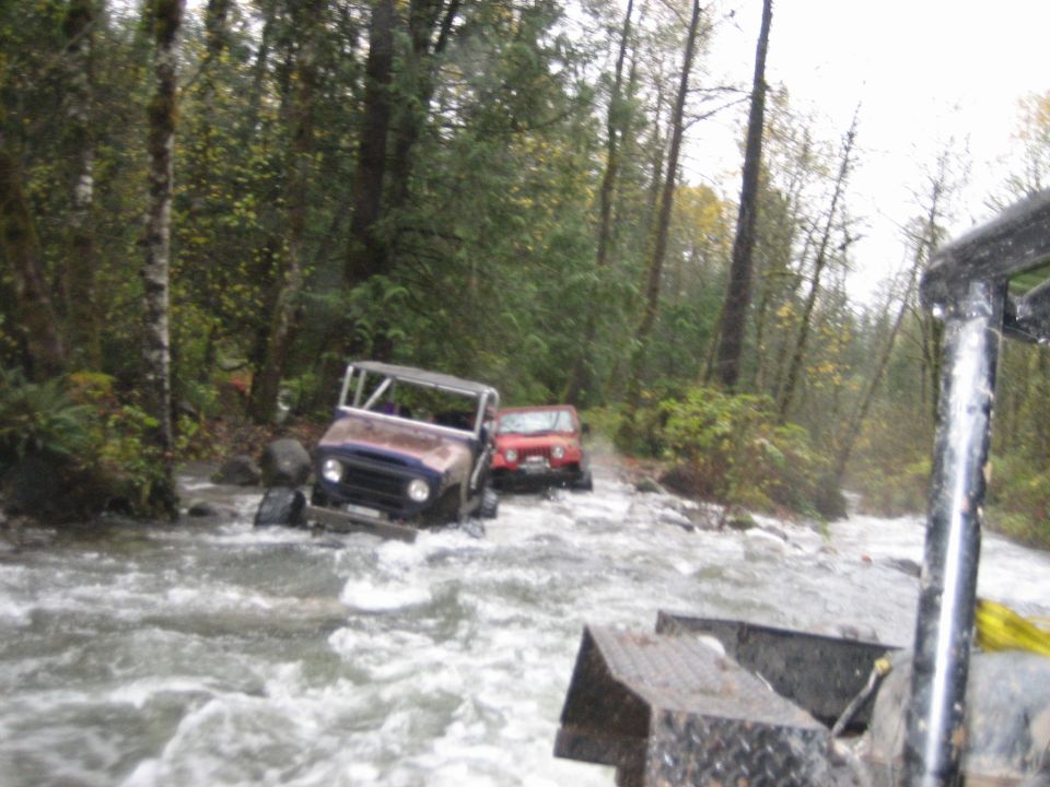 Two off-road vehicles driving through a river with trees on either side