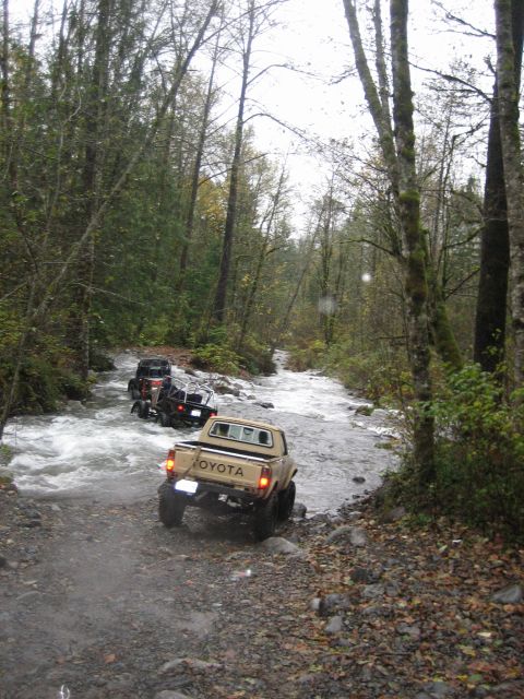 1990 Ford Bronco - Multiple off-road vehicles navigate a rocky riverbed in a wooded area.